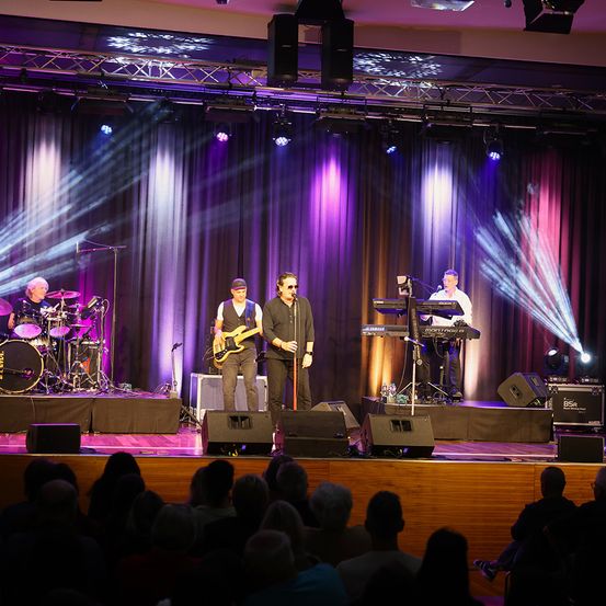 Eine Band spielt auf der Bühne mit lila und rosa Beleuchtung. Der Leadsänger spielt das Keyboard, während zwei Gitarristen und ein Schlagzeuger ihn begleiten. Das Publikum sitzt vor der Bühne.