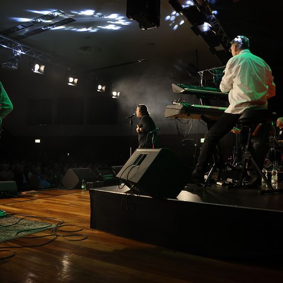 Eine Band tritt auf der Bühne auf, mit einem Sänger, der singt, und einem Keyboarder, der spielt. Die Bühne ist gut beleuchtet mit Scheinwerfern, und Lautsprecher sind in der Umgebung positioniert. Das Publikum sitzt darunter.