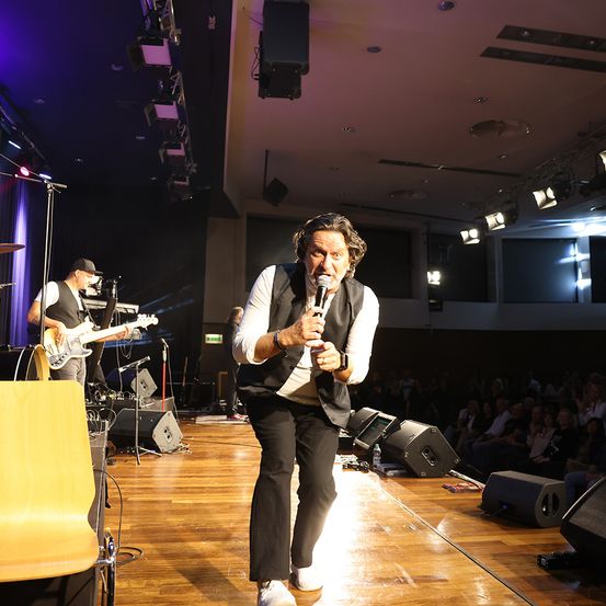 Ein Mann auf der Bühne singt in ein Mikrofon, während eine Band hinter ihm spielt. Die Bühne hat einen Holzboden und ist hell beleuchtet. Das Publikum sitzt vor der Bühne.