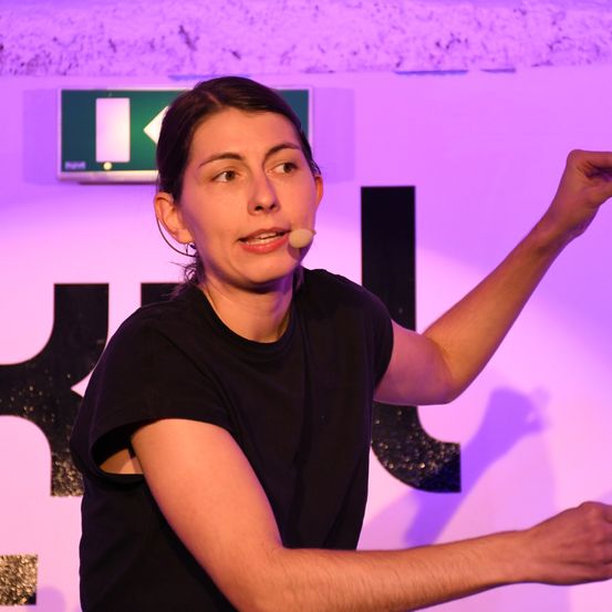 Eine Frau in einem schwarzen T-Shirt spricht in ein Mikrofon, während sie ihre rechte Hand hebt. Sie steht vor einer Wand mit einem lila-schwarzen Design.