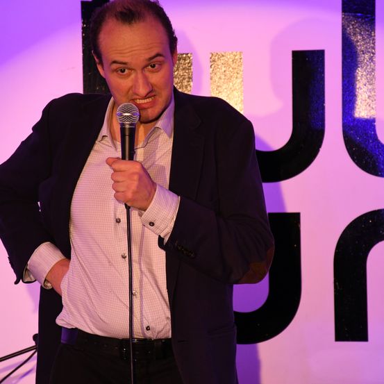 Ein Mann steht auf der Bühne, ein Mikrofon in der Hand, gekleidet in einen dunklen Anzug und ein weißes Hemd, vor einem Hintergrund mit großen Buchstaben.