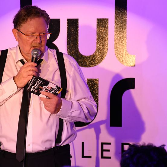 Ein Mann steht auf der Bühne und hält ein Mikrofon und ein Büchlein. Er trägt ein weißes Hemd, eine schwarze Krawatte und schwarze Hosenträger. Der Hintergrund ist lila und gold mit dem Wort 'KUL' in großen Buchstaben.