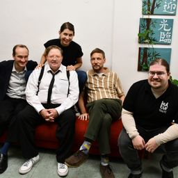 Fünf Personen sitzen auf einer roten Couch in einem Innenraum. Ein Mann auf der rechten Seite trägt eine Brille. Der Mann auf der linken Seite trägt eine Krawatte und ein Paar Turnschuhe. Hinter ihnen hängen drei Gemälde an der Wand.