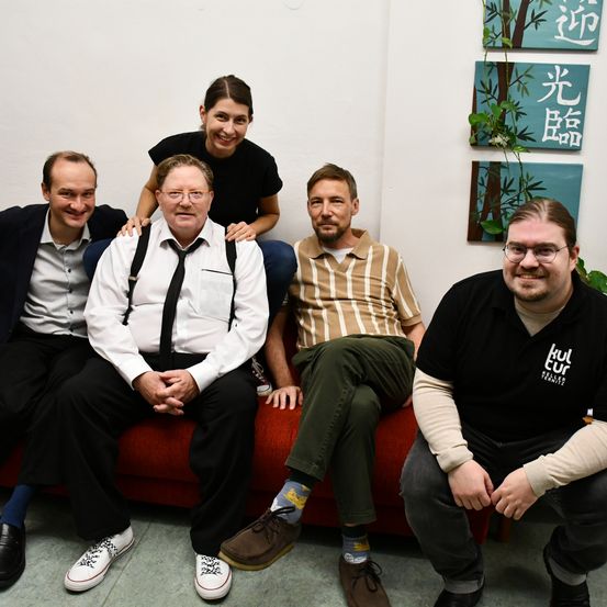 Fünf Personen sitzen auf einer roten Couch in einem Innenraum. Ein Mann auf der rechten Seite trägt eine Brille. Der Mann auf der linken Seite trägt eine Krawatte und ein Paar Turnschuhe. Hinter ihnen hängen drei Gemälde an der Wand.