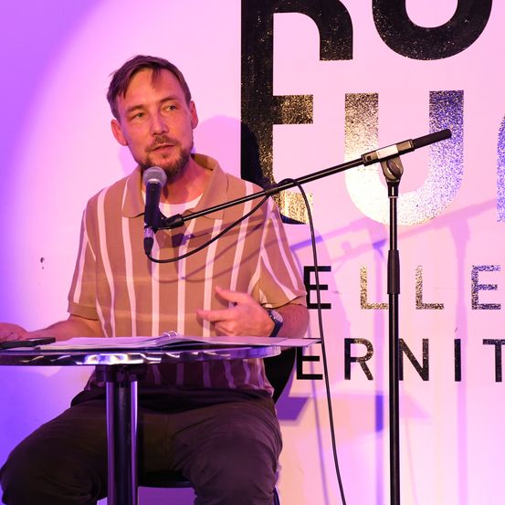 Ein Mann spricht in ein Mikrofon bei einer Konferenz mit einer lila Wand und großen schwarzen Buchstaben hinter ihm.