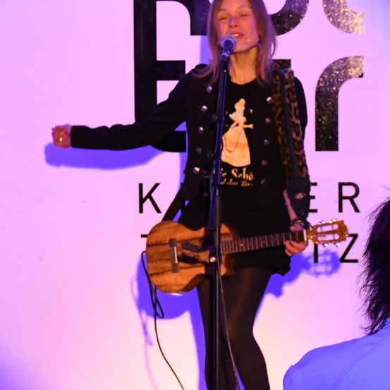 Eine Frau in einer schwarzen Jacke mit einem Disney-Charakter darauf singt in ein Mikrofon auf der Bühne.