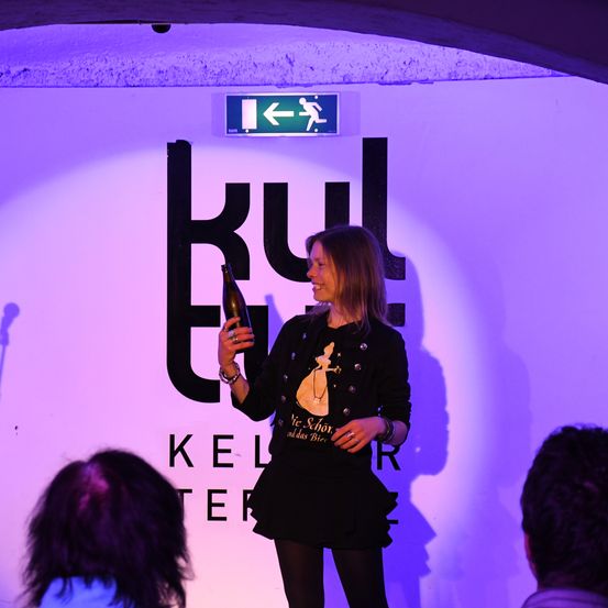 Eine Frau hält eine Flasche vor einem Schild mit der Aufschrift 'KELTER' und 'KUL' auf einem lila Hintergrund. Das Bild hat ein Schild über ihr mit einem Pfeil nach links.