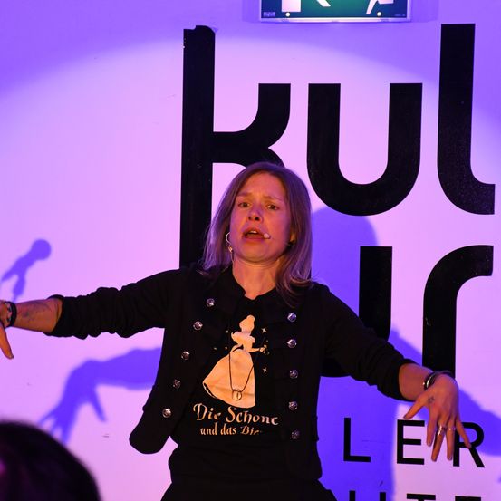 Eine Frau in einem schwarzen Jacken mit einem Cinderella-Aufdruck spricht in ein Mikrofon auf einer Bühne mit lila Hintergrund.