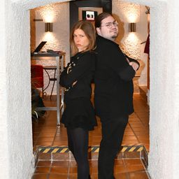 Ein Mann und eine Frau in Schwarz stehen mit verschränkten Armen in einem Bogengang, mit einem Tisch und einem roten Stuhl im Hintergrund.