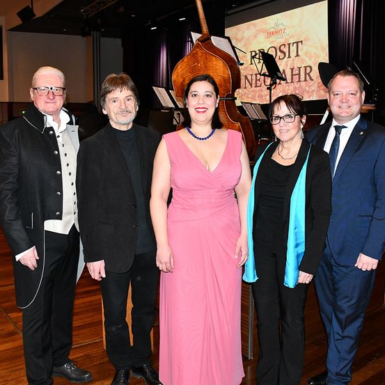 Fünf Menschen stehen lächelnd nebeneinander in einem Raum mit Holzboden. Sie befinden sich wahrscheinlich bei einer formellen Veranstaltung. Eine Frau in einem rosa Kleid steht in der Mitte, mit einem Cello und einem Notenständer hinter ihr.
