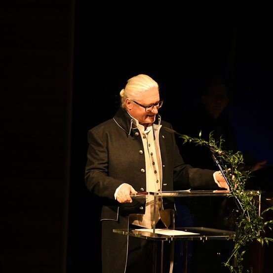 Ein älterer Mann mit Brille und schwarzem Mantel steht an einem Podium mit Mikrofon, hält ein Stück Papier in der Hand und lächelt.