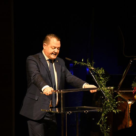Ein Mann in Anzug und Krawatte steht an einem Rednerpult und hält ein Messer, möglicherweise um ein Band zu schneiden. Ein Mikrofon ist auf dem Pult positioniert, und eine grüne Pflanze ist in der Nähe arrangiert. Die Umgebung scheint ein formelles Ereignis zu sein.
