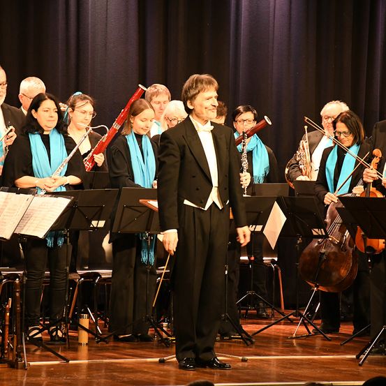 Eine Gruppe von Musikern in Schwarz und Blau spielt auf einer Bühne mit schwarzem Vorhang. Sie spielen verschiedene Instrumente wie Flöten, Celli und Klarinetten.