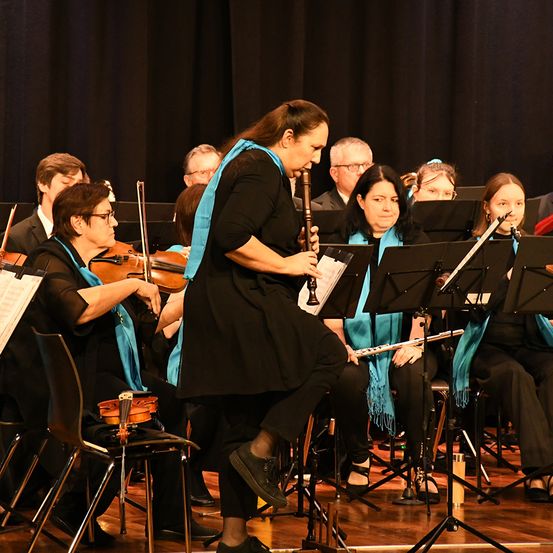 Eine Gruppe von Musikern, gekleidet in Schwarz und Blau, tritt auf der Bühne auf. Die Frau in der Mitte spielt Flöte. Violinen stehen auf Stühlen vor den Musikern. Das Publikum sitzt hinter ihnen.