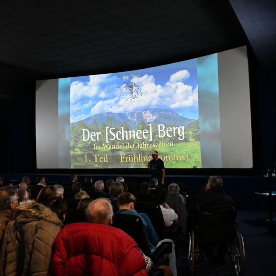 Ein Publikum sitzt in Stühlen und schaut sich eine große Leinwand mit einem Film namens 'Der Schneel Berg' an. Ein Mann steht vor der Leinwand.