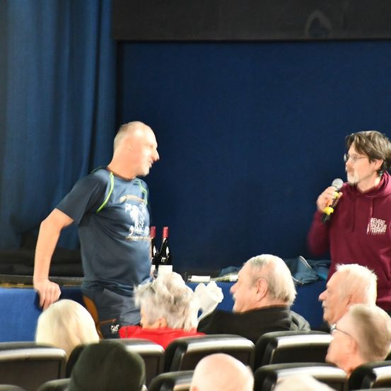 Zwei Männer stehen auf der Bühne, einer mit einem Mikrofon, sie sprechen eine Zuhörerschaft an. Flaschen stehen auf dem Tisch. Zuschauer sitzen in Reihen, einige mit Brille.