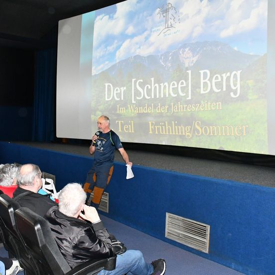 Ein Mann steht mit einem Mikrofon auf der Bühne und hält wahrscheinlich eine Präsentation. Hinter ihm zeigt eine große Leinwand eine landschaftliche Berglandschaft. Mehrere Personen sitzen vor der Bühne.