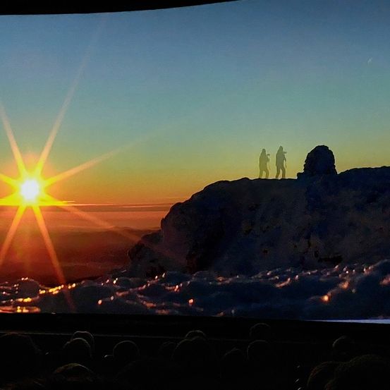 Ein atemberaubender Sonnenuntergang über einem verschneiten Berggipfel mit zwei Silhouetten von Menschen, die oben stehen. Der Himmel ist eine Mischung aus orange, pink und blauen Farbtönen.