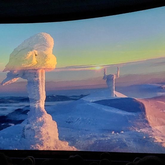 Eine verschneite Landschaft zeigt eine einzigartige Schneeformation, die einem Kopf ähnelt. Dahinter steht eine weitere Schneefigur. Der Himmel ist blau mit Anflügen von Grün und Gelb, und Berge sind in der Ferne sichtbar.