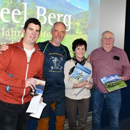 Vier Personen posieren vor einem Bildschirm mit Bergkulisse, jeder ein Buch in der Hand und lächelnd.