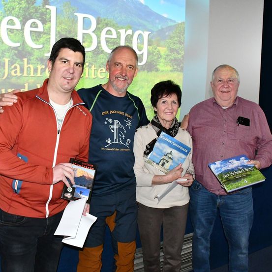 Vier Personen posieren vor einem Bildschirm mit Bergkulisse, jeder ein Buch in der Hand und lächelnd.