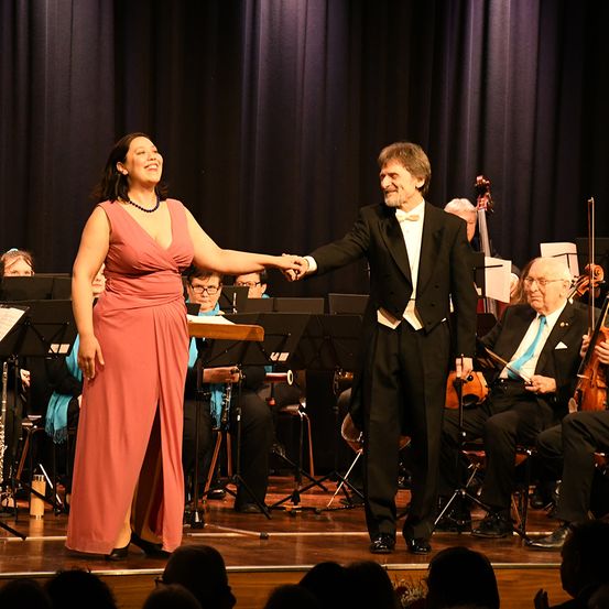 Ein Mann und eine Frau stehen lächelnd und Hand in Hand auf der Bühne. Dahinter spielen Musiker verschiedene Instrumente. Das Publikum beobachtet vom ersten Rang aus.