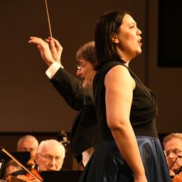 Eine Sängerin steht vor einem Orchester und singt mit offenem Mund. Der Dirigent hinter ihr hebt seinen Taktstock. Die Orchestermitglieder spielen ihre Instrumente.