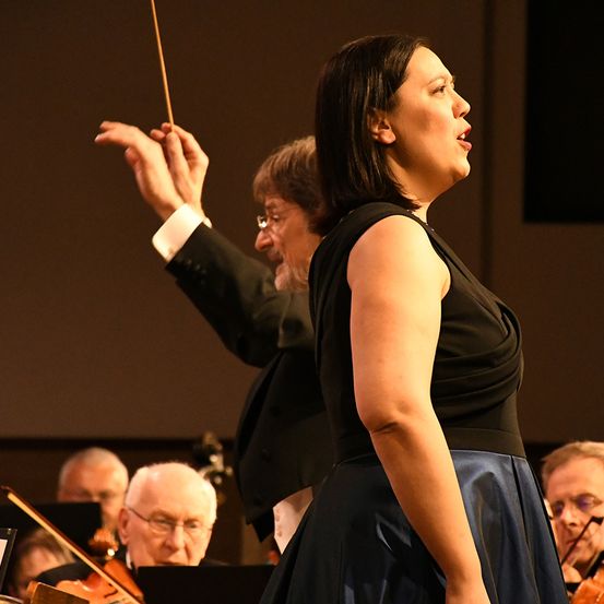 Eine Sängerin steht vor einem Orchester und singt mit offenem Mund. Der Dirigent hinter ihr hebt seinen Taktstock. Die Orchestermitglieder spielen ihre Instrumente.
