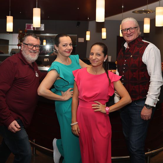 Vier Personen stehen lächelnd vor einer Bartheke für ein Foto. Die beiden Frauen tragen Kleider und die Männer Freizeitkleidung.