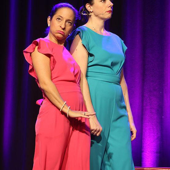 Zwei Frauen stehen auf einer Bühne, eine in einem rosa Kleid und die andere in einem türkisen Kleid. Beide schauen nach vorne, möglicherweise bei einer Aufführung. Die Frau in Rosa hat einen ernsten Ausdruck, während die andere ein Mikrofon trägt. Der Hintergrund ist ein violetter Vorhang.