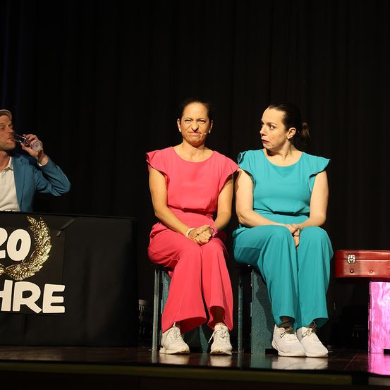 Zwei Frauen in rosa und blauen Outfits sitzen auf der Bühne und blicken sich an. Ein Mann trinkt hinter ihnen aus einem Becher. Ein Tisch mit einer goldenen Girlande und der Zahl '20' im Hintergrund.