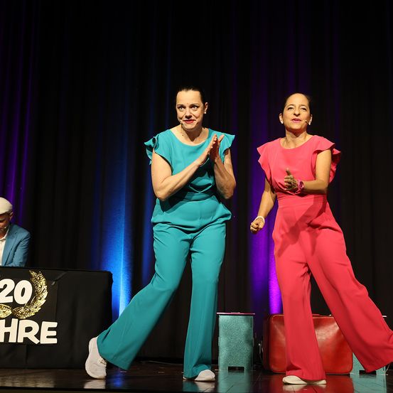 Zwei Frauen tanzen auf der Bühne, eine in Grün und die andere in Rosa. Hinter ihnen ein schwarzer Hintergrund mit '20 Jahre' Text. Ein Mann sitzt an einem Schreibtisch mit Mikrofon.