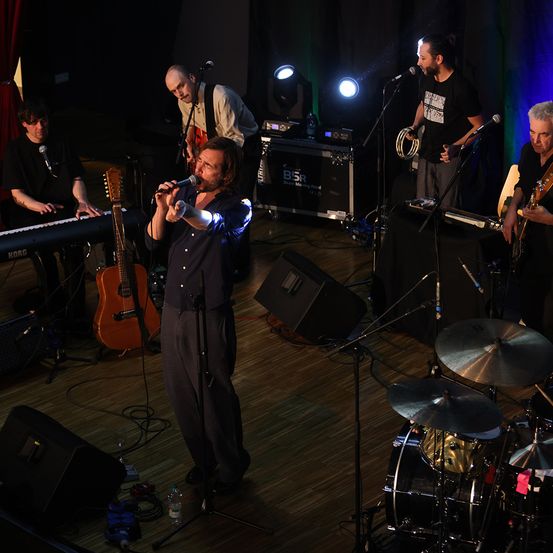Eine Band spielt auf der Bühne. Ein Mann singt in ein Mikrofon, während ein anderer Gitarre spielt. Die Band besteht aus einem Pianisten, einem Schlagzeuger und einem Perkussionisten. Scheinwerfer und Lautsprecher sind auf der Bühne aufgebaut.