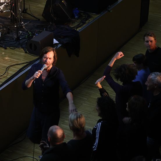 Ein Mann in einem dunklen Hemd singt in ein Mikrofon auf der Bühne mit einer Band. Ein Publikum aus Menschen steht um ihn herum.