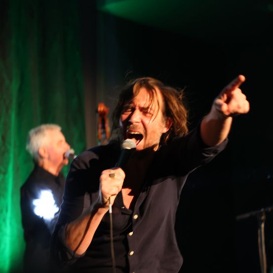 Ein Mann singt leidenschaftlich in ein Mikrofon auf der Bühne, während ein anderer Musiker Cello spielt.