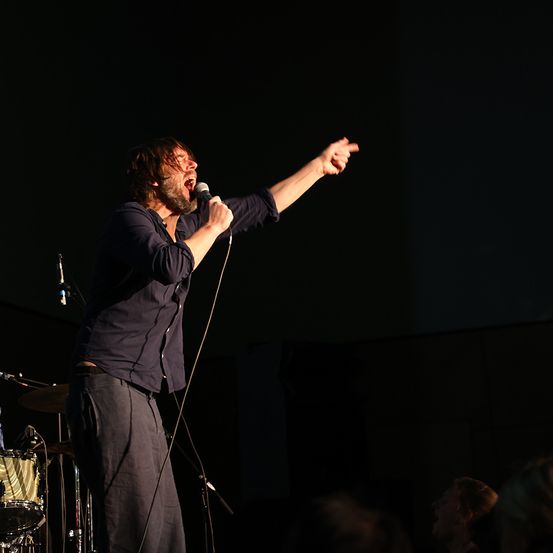 Ein Mann singt leidenschaftlich in ein Mikrofon auf der Bühne und gestikuliert mit der rechten Hand. Dahinter ist ein Schlagzeug zu sehen, und ein Mikrofonstativ steht bereit. Der Hintergrund ist dunkel.