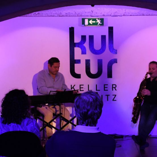 Zwei Musiker treten in einem Veranstaltungsort namens Kellerwitz auf. Ein Mann spielt Saxophon, während der andere ein Keyboard spielt. Einige Leute beobachten sie aus dem Publikum.