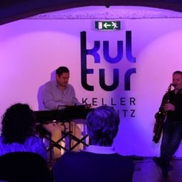 Zwei Musiker treten in einem Veranstaltungsort namens Kellerwitz auf. Ein Mann spielt Saxophon, während der andere ein Keyboard spielt. Einige Leute beobachten sie aus dem Publikum.