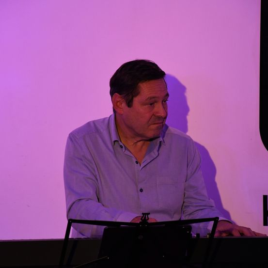 Ein Mann in einem blauen Hemd sitzt und spielt Klavier oder Keyboard, mit einem lila Hintergrund hinter ihm. Er schaut zur Seite.