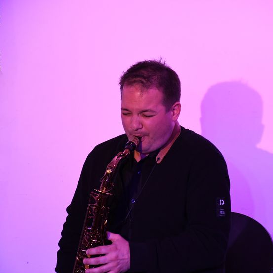 Ein Mann in einem schwarzen Pullover spielt Saxophon unter lila Licht. Er konzentriert sich auf seine Darbietung und steht vor einer lila Wand.