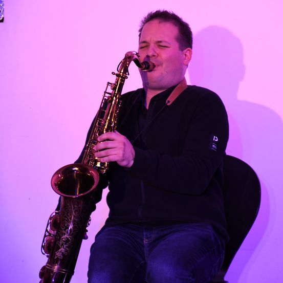 Ein Mann sitzt auf einem Stuhl und spielt Saxophon. Er trägt ein schwarzes Langarmhemd und blaue Jeans. Der Hintergrund ist lila.