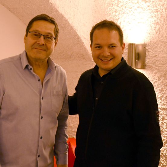 Zwei Männer stehen eng nebeneinander und lächeln. Der Mann auf der linken Seite trägt ein hellblaues Hemd. Der Mann auf der rechten Seite trägt eine dunkle Jacke. Hinter ihnen ist eine gebogene Wand mit einer Beleuchtungseinrichtung.