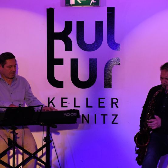 Zwei Männer treten auf einer Bühne unter einer lila Beleuchtung auf. Einer spielt Klavier, der andere Saxophon. An der Wand hinter ihnen steht das Wort 'Kultur' in großen Buchstaben.