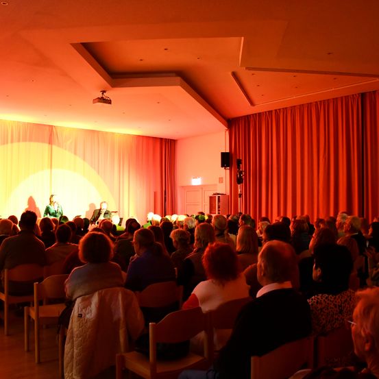 Ein Theater mit rotem Vorhang und Scheinwerfer auf der Bühne mit zwei Personen. Das Publikum sitzt auf Stühlen.