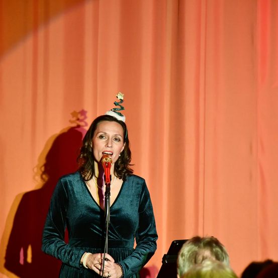 Eine Frau mit einem Weihnachtsbaum-Kopfband singt in ein Mikrofon auf einer Bühne mit einem orangefarbenen Vorhang im Hintergrund.