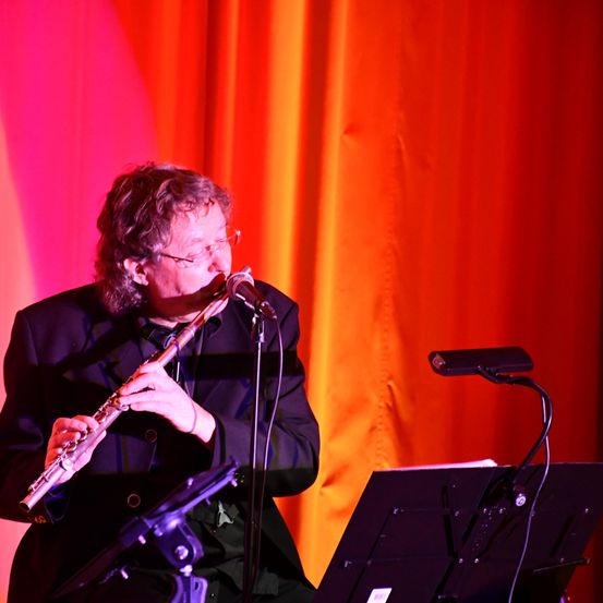 Ein Mann mit Brille und Anzug spielt Flöte vor einem Mikrofon. Die Bühne hat einen roten Vorhang und einen schwarzen Notenständer.