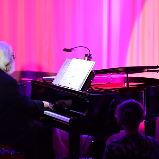 Ein älterer Mann spielt Klavier, davor ein Notenständer, beleuchtet von rosa Licht. Ein junger Mensch sitzt davor.