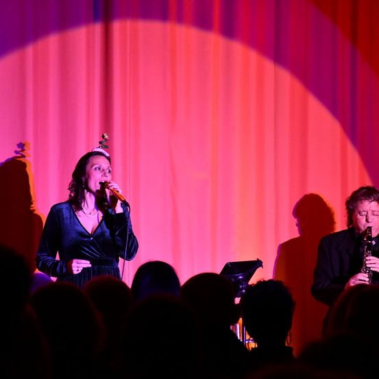 Eine Frau singt auf der Bühne mit einem Mikrofon vor einem Publikum. Ein Mann spielt Klarinette. Sie werden von einem Scheinwerfer auf einem roten Vorhang beleuchtet.