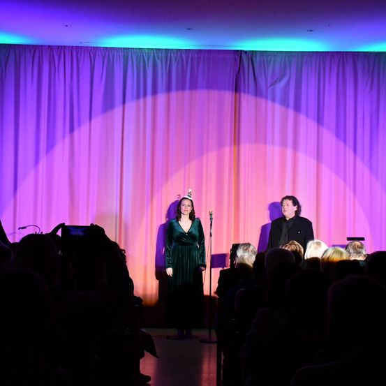 Eine Frau in einem grünen Kleid mit einer Party-Mütze steht mit einem Mann auf der Bühne und schaut das Publikum an. Die Bühne ist mit lila und orangefarbenen Lichtern beleuchtet, und das Publikum sitzt vor ihnen.