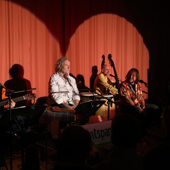 Eine Band spielt auf der Bühne mit rotem Hintergrund, bestehend aus einem Gitarristen, einem Cellisten, einem Perkussionisten und einem Sänger. Der Gitarrist und der Cellist sitzen, während der Sänger steht.